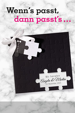 Schwarze Einladungskarte zur Hochzeit, an die mit einer weißen Schleife zwei Puzzleteile angeknüpft sind.