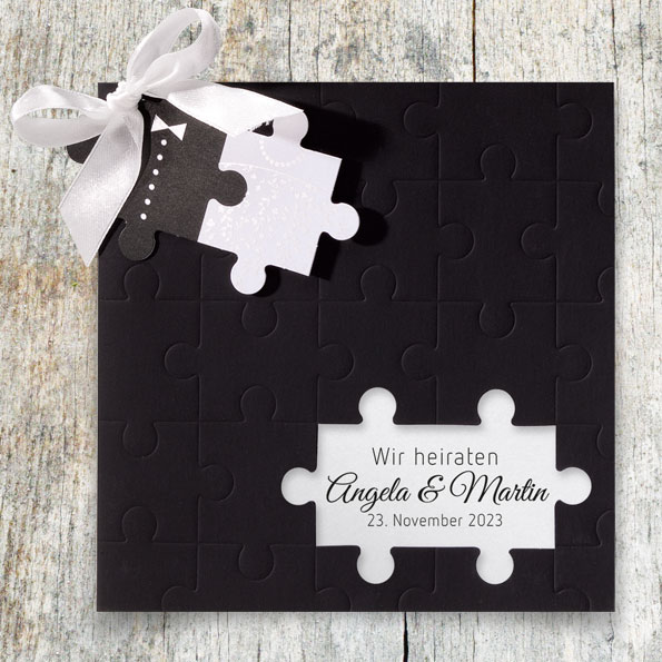 Schwarze Einladungskarte zur Hochzeit, an die mit einer weißen Schleife zwei Puzzleteile angeknüpft sind.