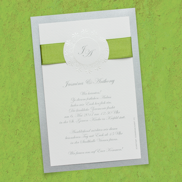Weiße Hochzeitseinladungen, aufgeklebt auf silbernem, schillernden Trägerkarton, oben von einer grünen Schleife umfasst.