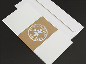 Eine unbedruckte Einladungskarte, umfasst von einer brunen Banderole, auf einem weißen Kuvert liegend.